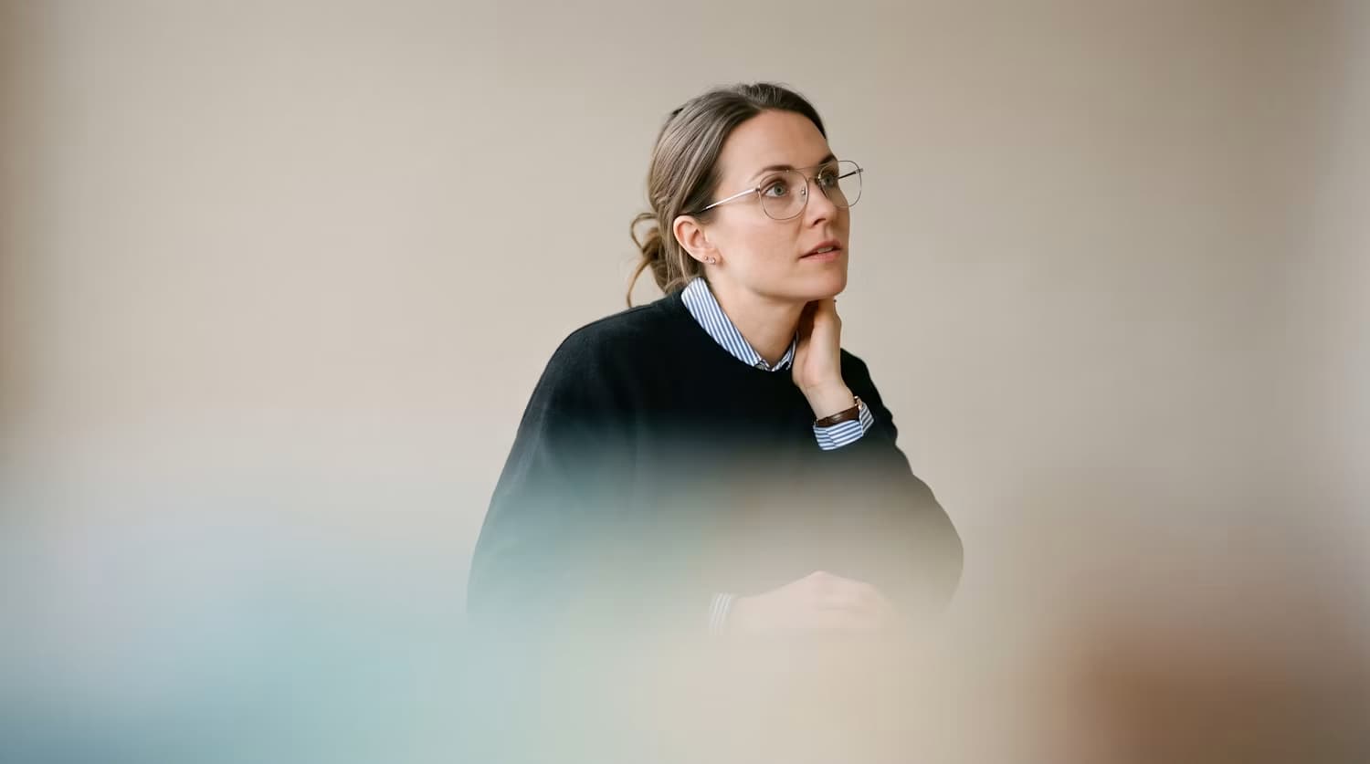 Une femme pensive, lunettes et pull sombre, le regard tourné vers le haut